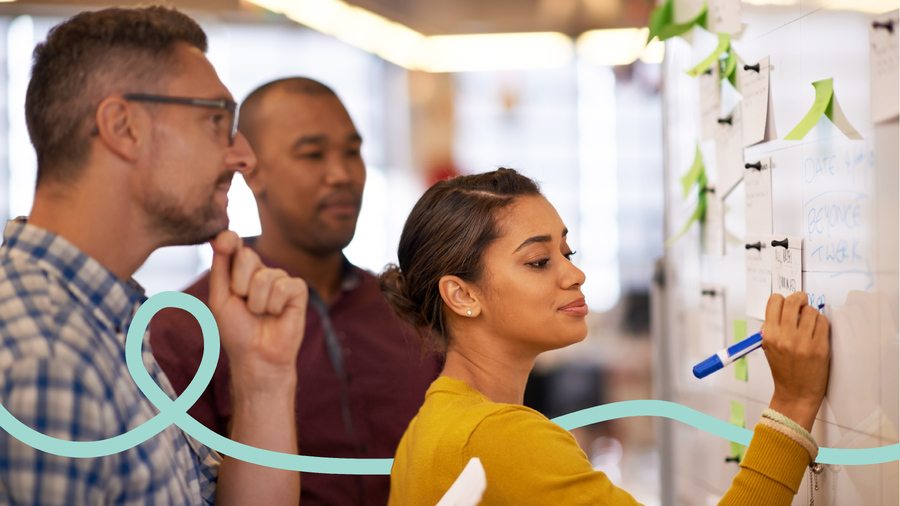 A diverse creative team collaborating in a bright office environment, with a woman writing ideas on a whiteboard alongside sticky notes while her colleagues look on thoughtfully during a brand naming brainstorming session.
